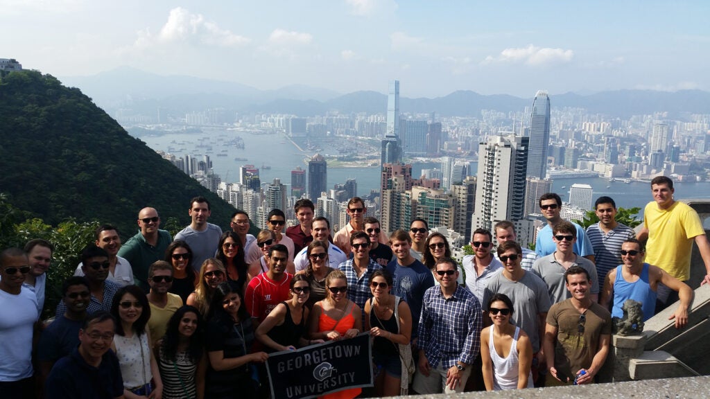 Georgetown MBA Students pose with the Hong Kong skyline in the background and the group is holding a flag that says Georgetown University